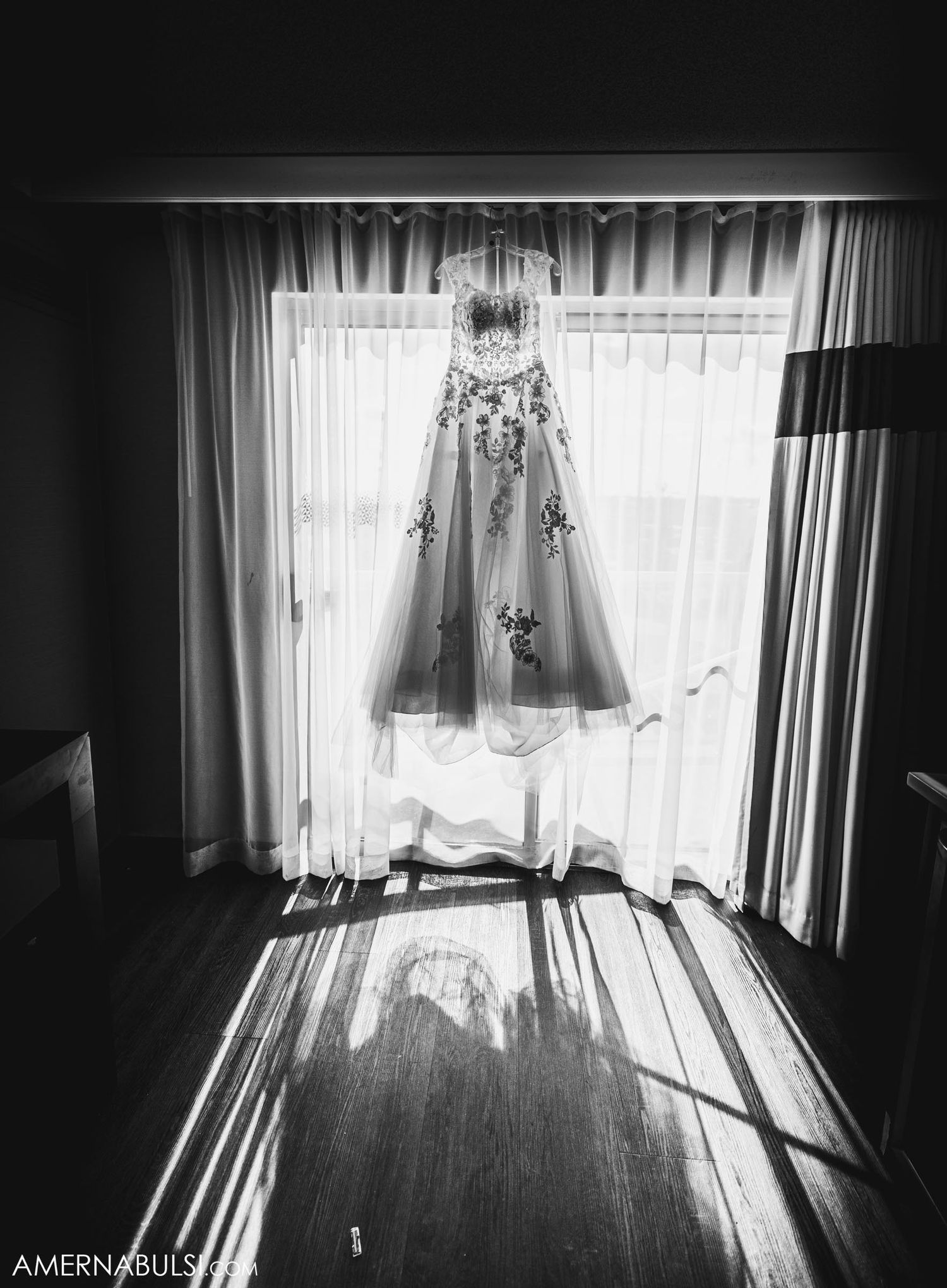 Black and white photo of wedding dress hanging in window with dramatic shadows.
