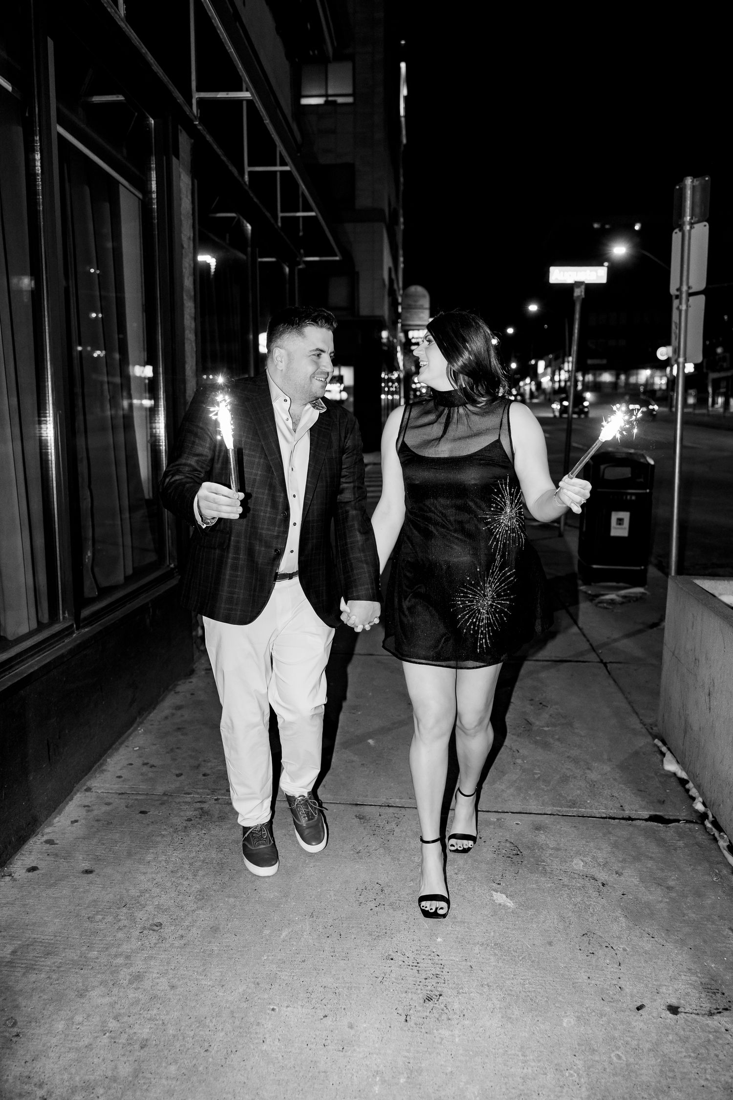 Black and white photo of a couple holding sparklers while walking hand in hand on a city sidewalk at night.