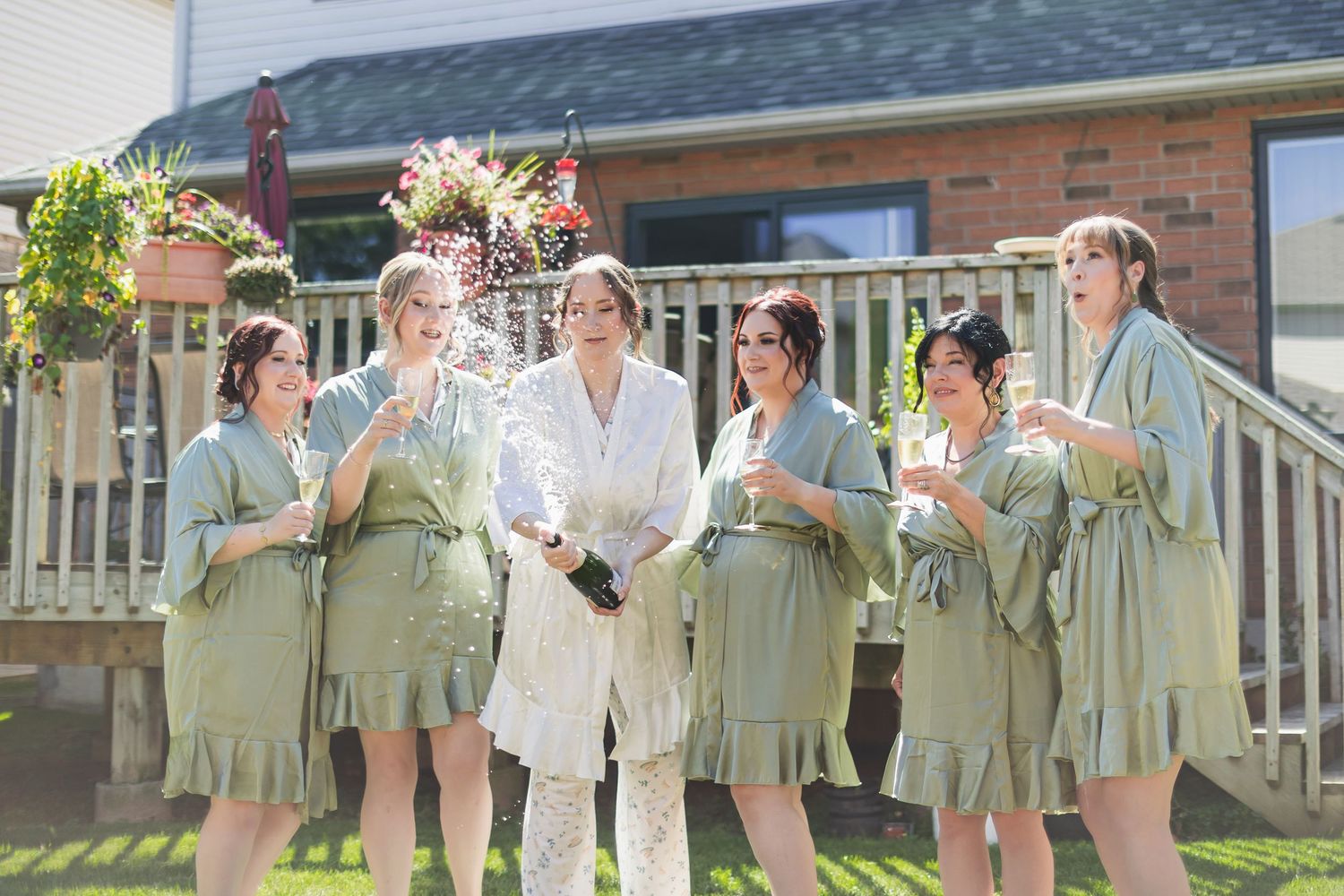 Bride and bridesmaids in sage robes pop champagne bottle outdoors on sunny wedding morning.