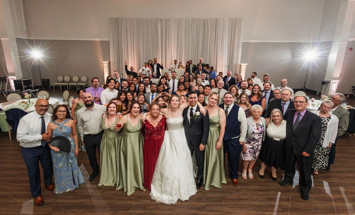 Large wedding group photo with bride in white gown, bridesmaids in green dresses, and guests gathered in elegant venue.