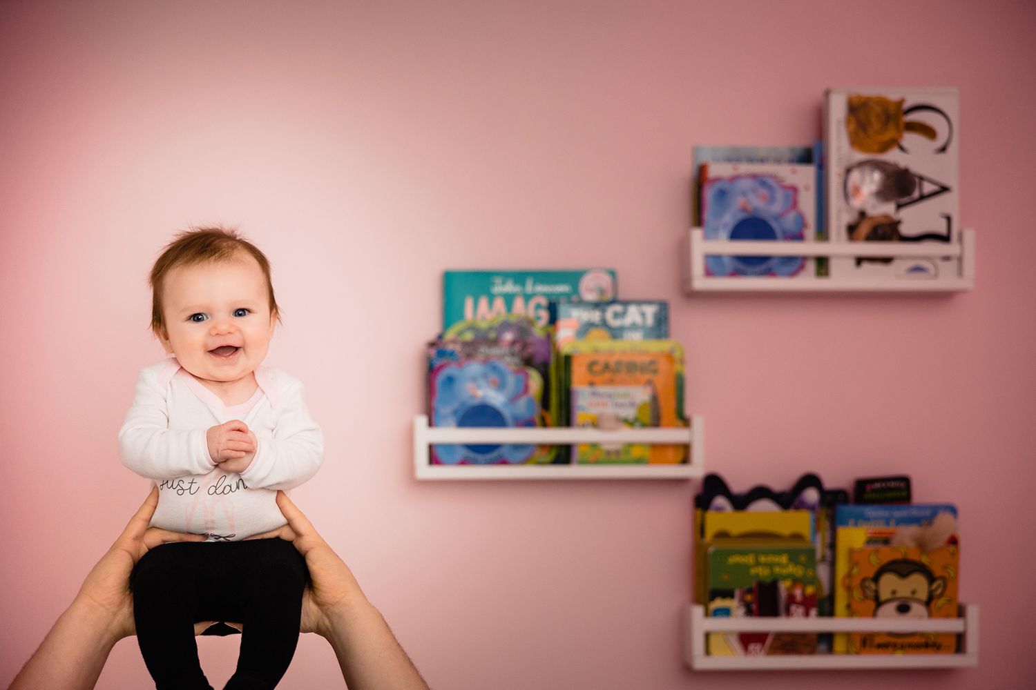 Baby sitting on raised hands against pink wall with floating bookshelves displaying children's books.