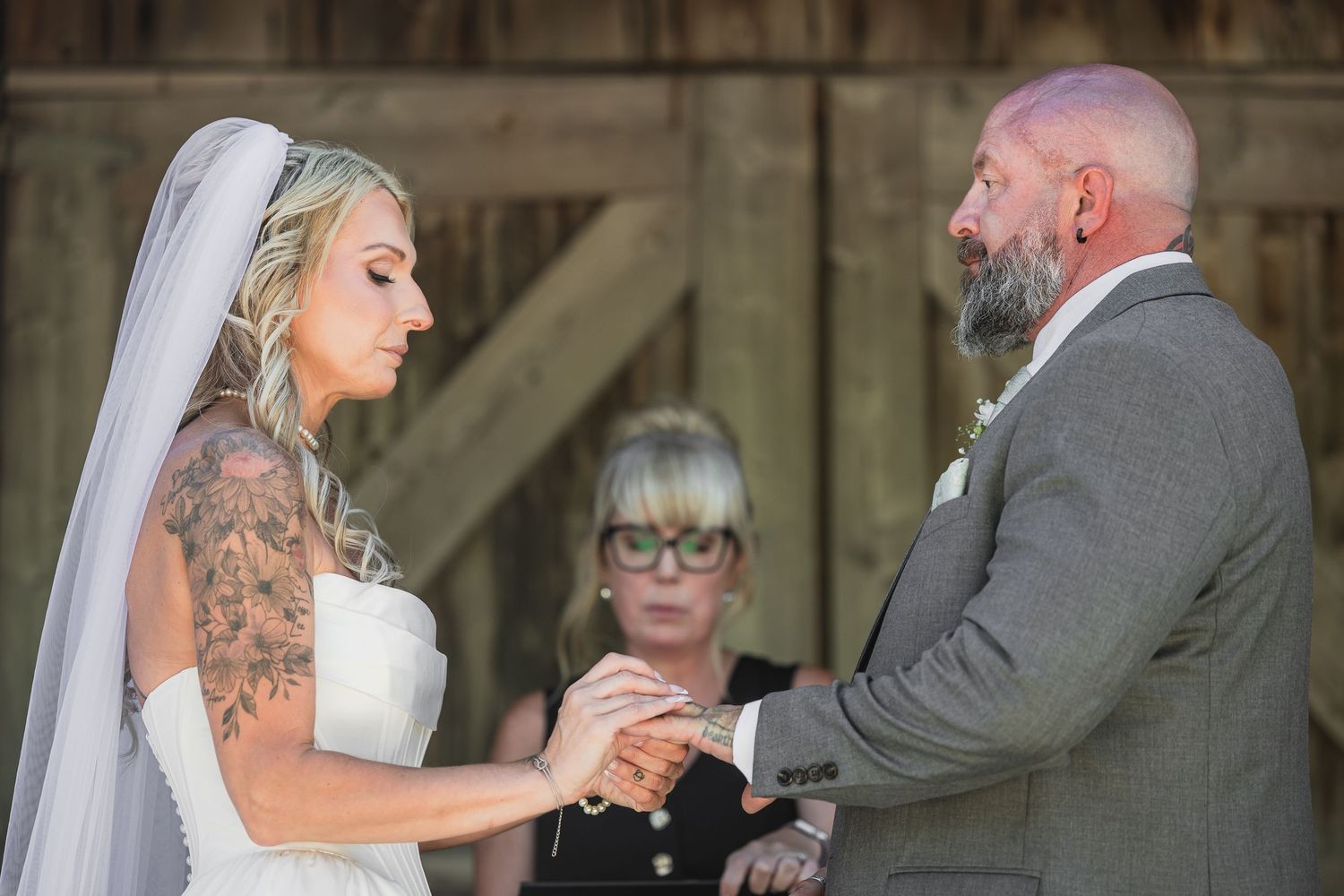 Tattooed bride exchanging rings with groom during intimate barn wedding ceremony with officiant in background.
