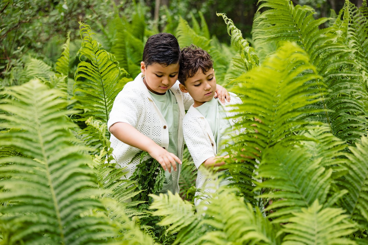Two young children play together in a lush garden filled with tall green ferns.
