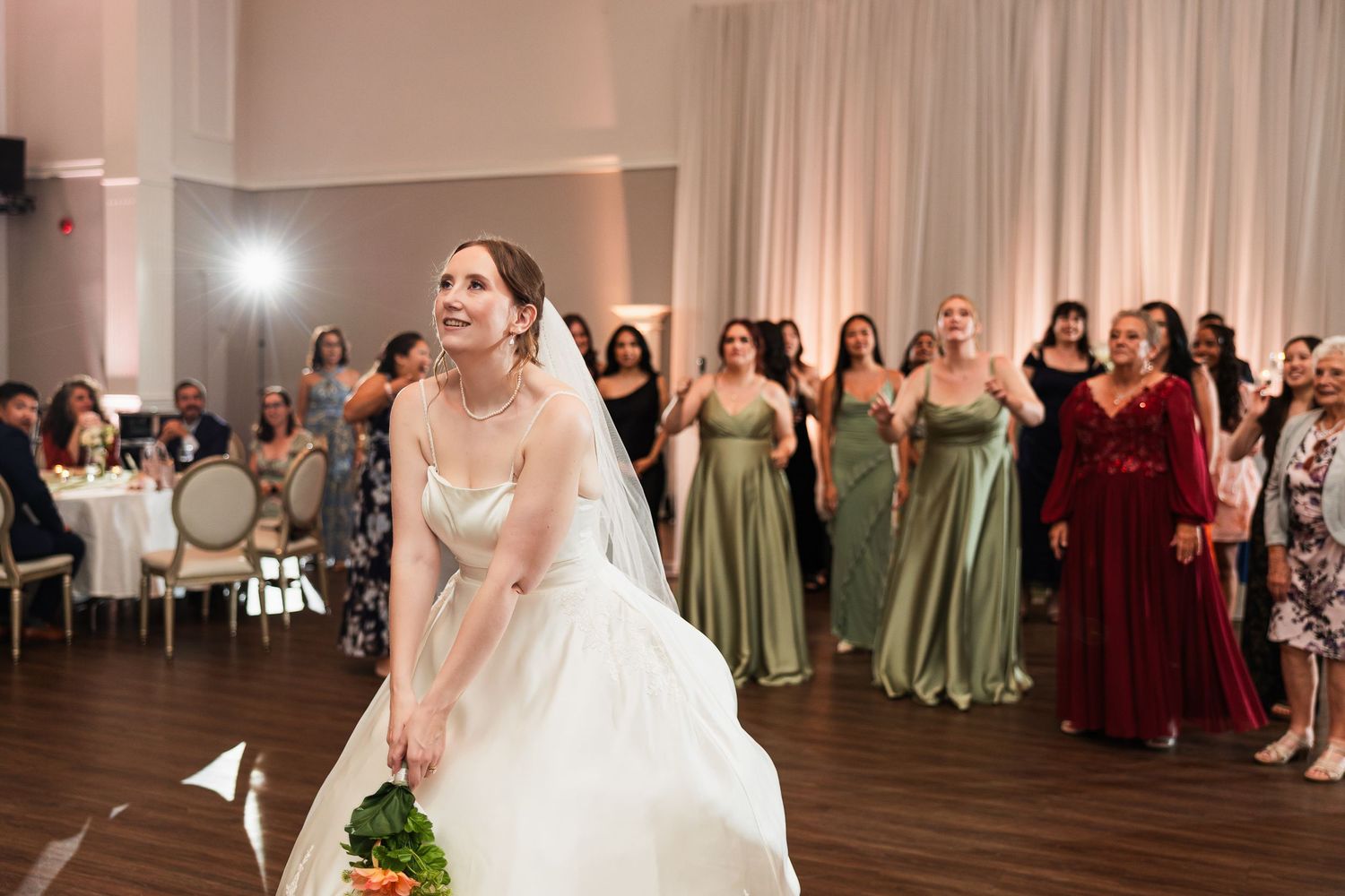 Bride in white gown tossing bouquet to bridesmaids in green dresses at elegant wedding reception.