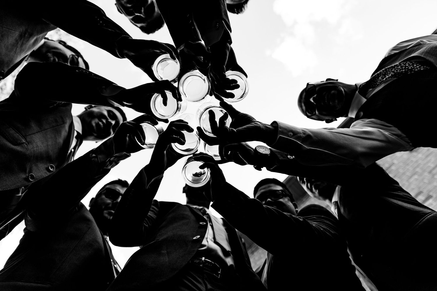 Black and white low angle shot of groomsmen toasting glasses together in a circle at wedding.