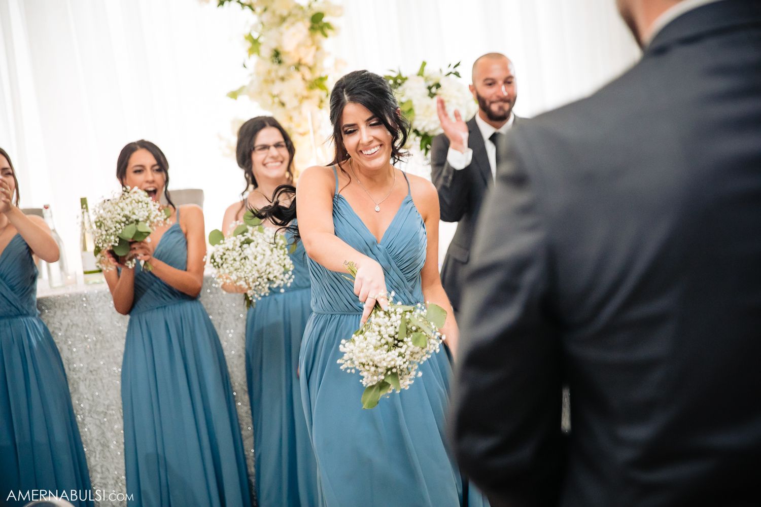 Bridesmaids in flowing dusty blue dresses holding white flower bouquets during ceremony.