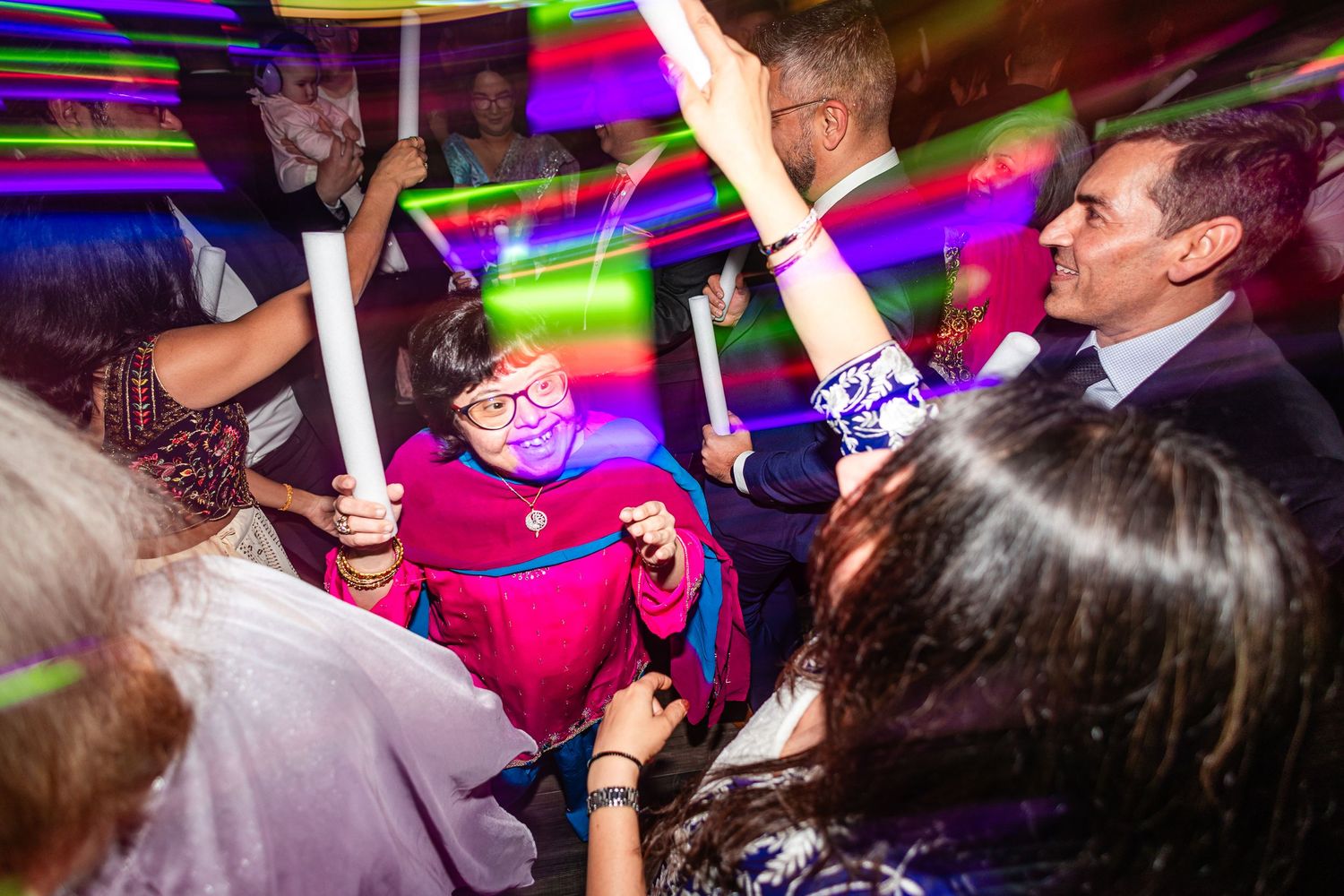 Partygoers wave glowing neon light sticks on a crowded dance floor with purple and green light trails.