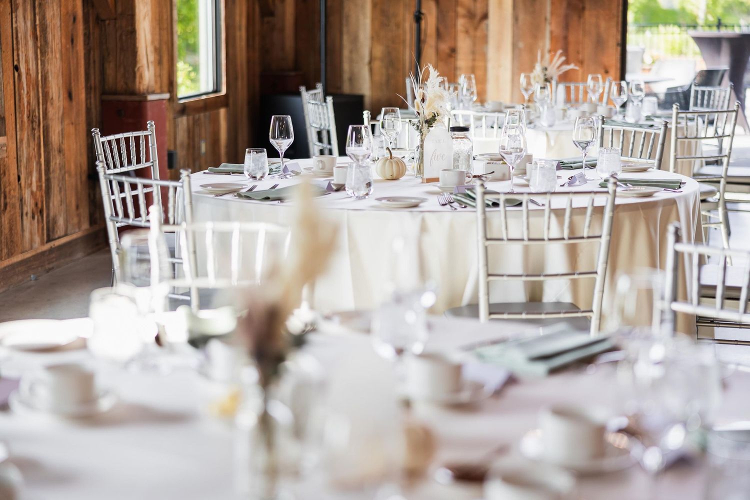 Rustic barn wedding reception setup with white linens and silver chiavari chairs in natural light.
