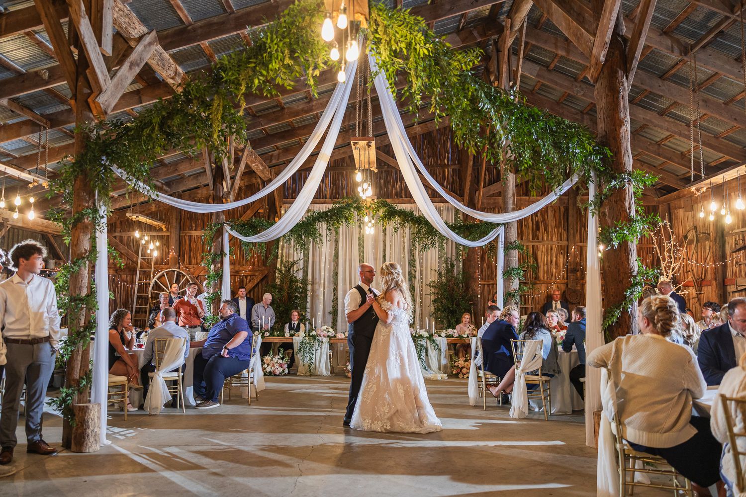 Wedding guests dance and celebrate in a rustic barn venue decorated with white fabric draping and twinkling lights.