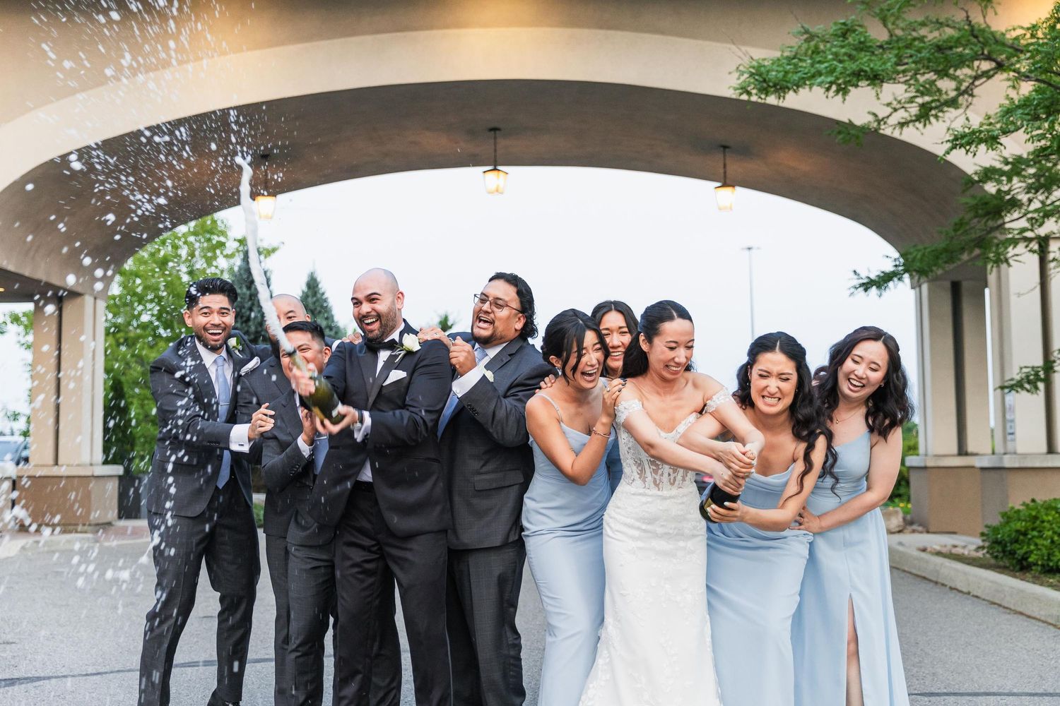 Wedding party celebrates with champagne spray under an architectural archway.