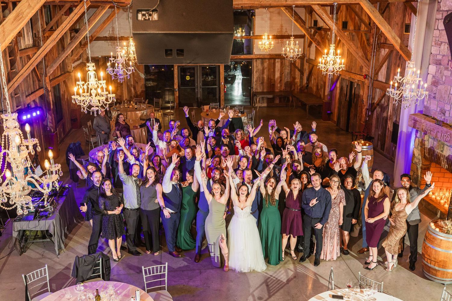 Large wedding party group photo in rustic barn venue with elegant chandeliers and purple lighting.