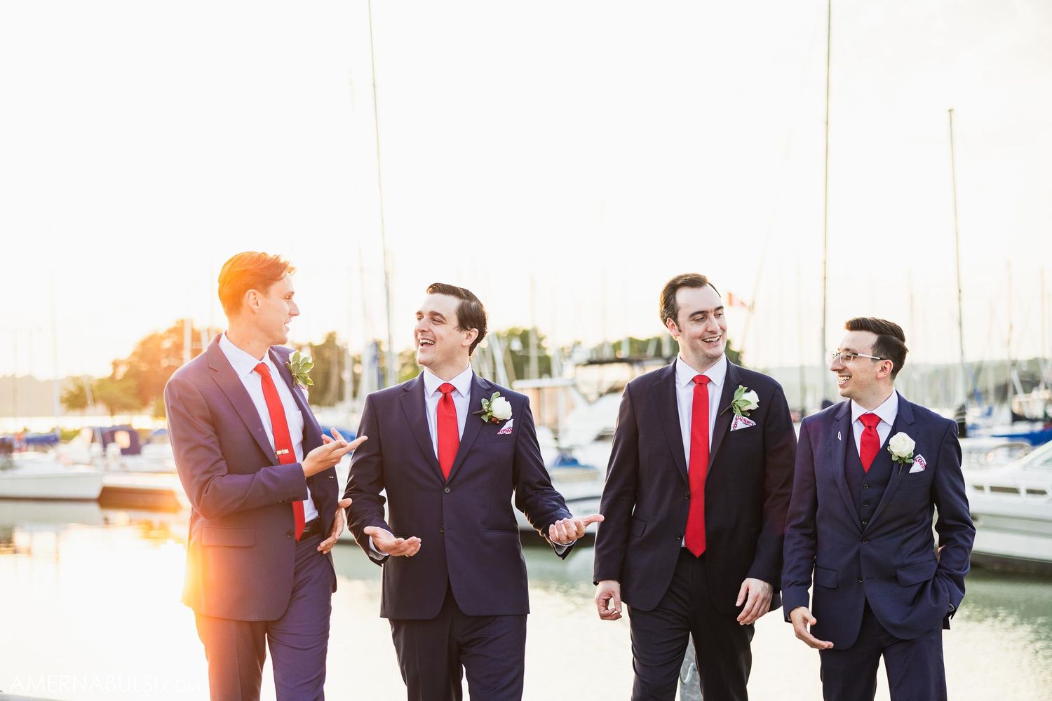 Groomsmen in navy suits with red ties chatting at sunset by the marina.
