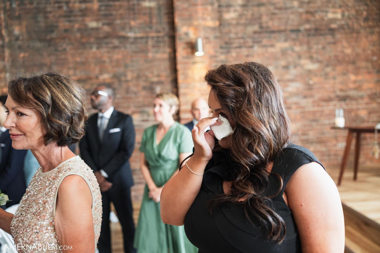 Wedding guests showing emotional reactions during ceremony in brick-walled venue.