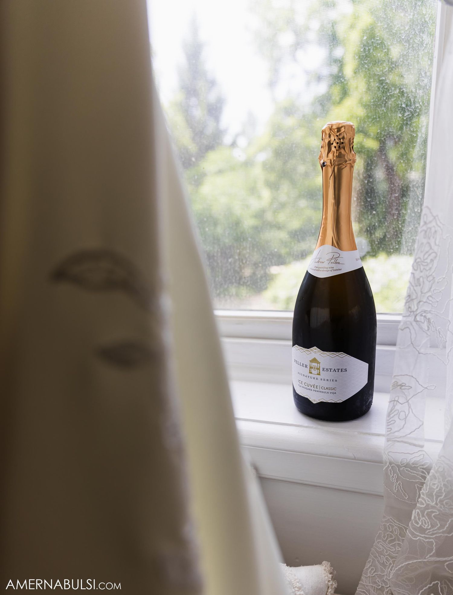 Bottle of champagne sitting on white windowsill with sheer curtains.