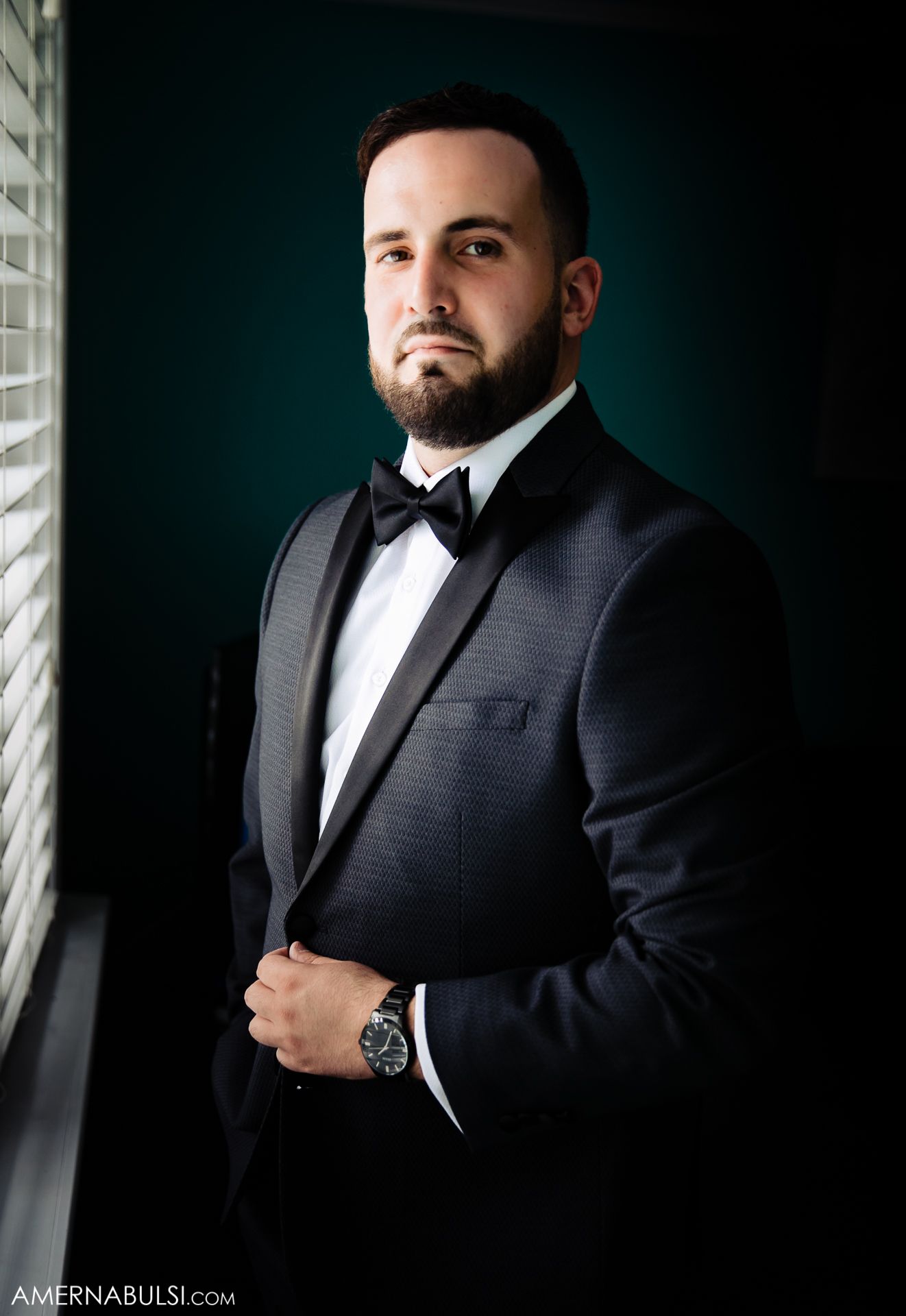 Professional studio portrait of someone wearing a black tuxedo and bow tie against a dark background.