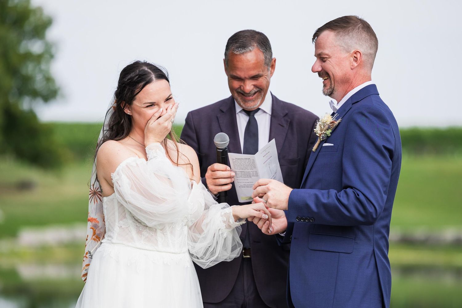 A touching wedding ceremony exchange takes place on a sunny day with a pond and green landscape behind.