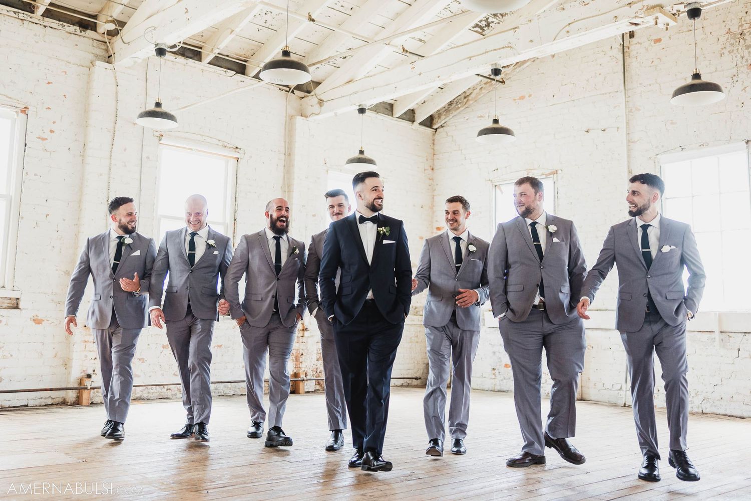Wedding party of men in grey suits and black tux pose casually in industrial white room with pendant lights.
