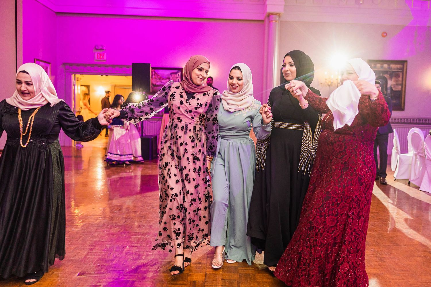 Women in elegant formal dresses dancing together at a wedding reception with pink lighting.
