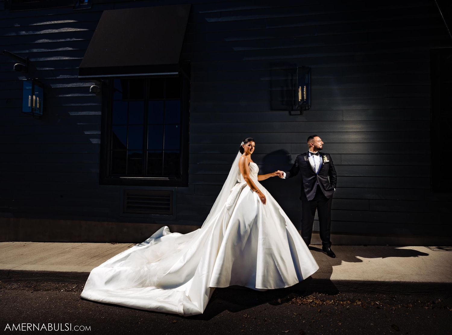A wedding couple poses dramatically against a dark building wall with a flowing white bridal gown.