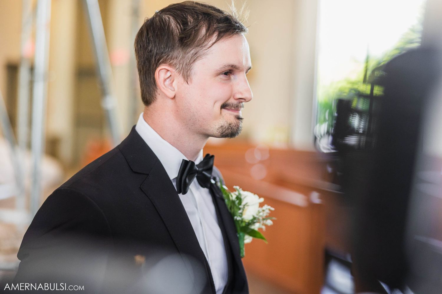 A groom wearing a black tuxedo and bowtie smiles during the wedding ceremony.
