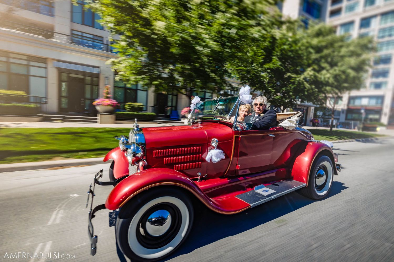 A shiny red vintage convertible car cruising down a tree-lined urban street.