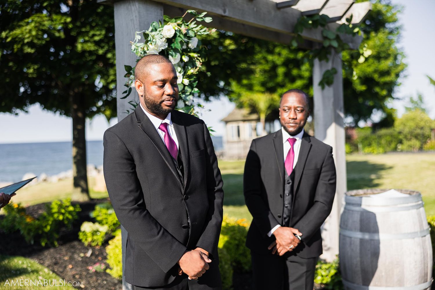 Wedding attendants in black suits and pink ties stand beneath a white pergola by the ocean.