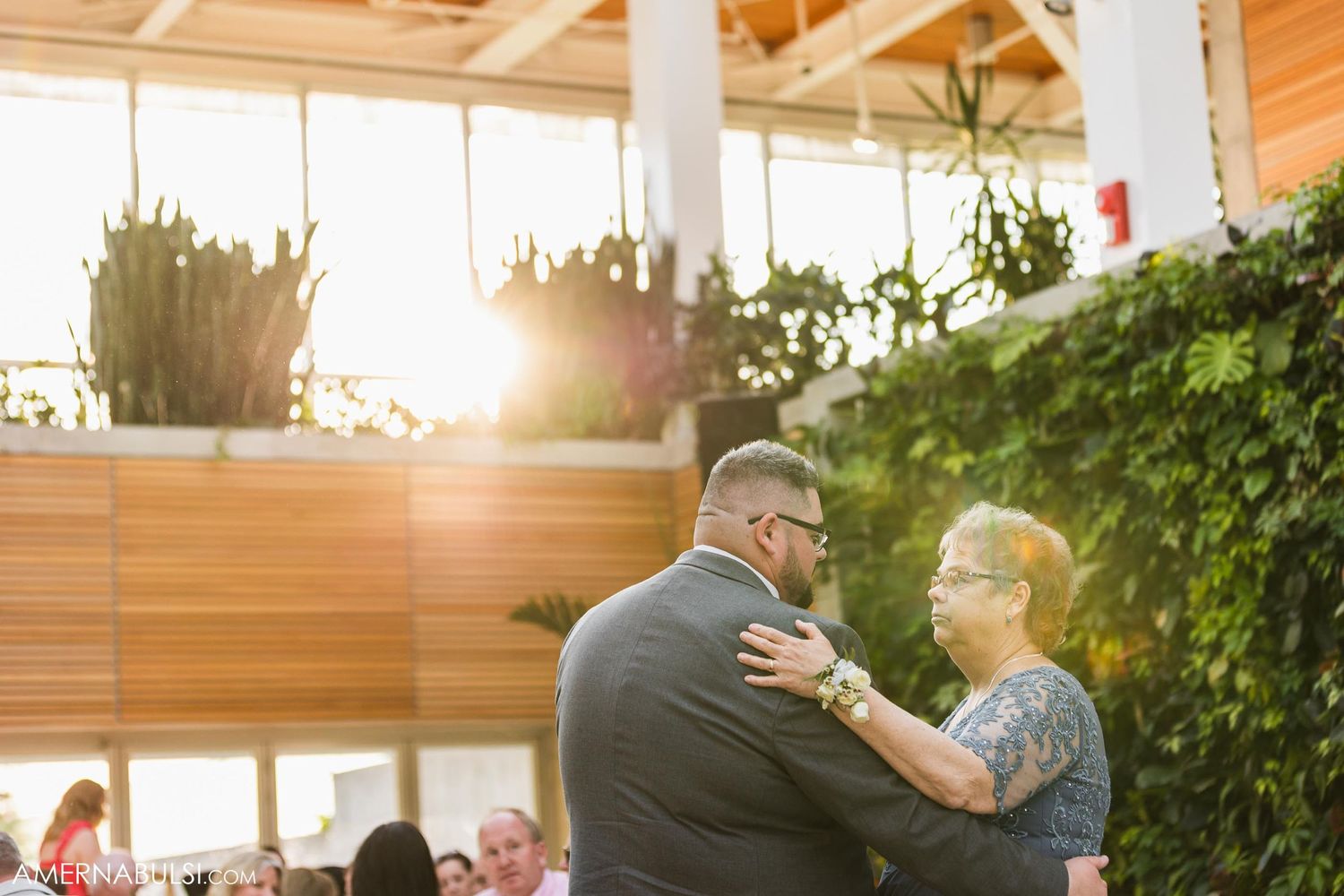 Two people share a tender dance moment at an outdoor reception with greenery and sunset lighting in the background.