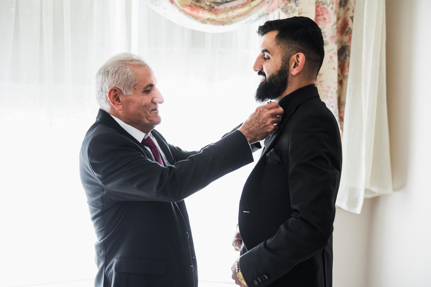 An older gentleman adjusts the tie of a younger man in formal attire by a window.
