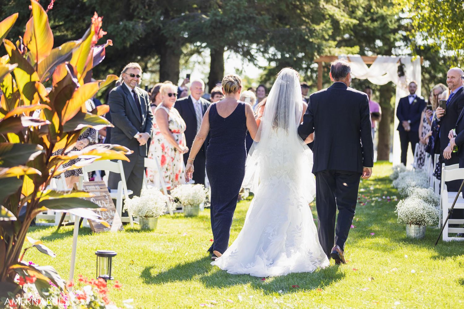 Wedding party walking down an outdoor aisle lined with white chairs on a bright sunny day.