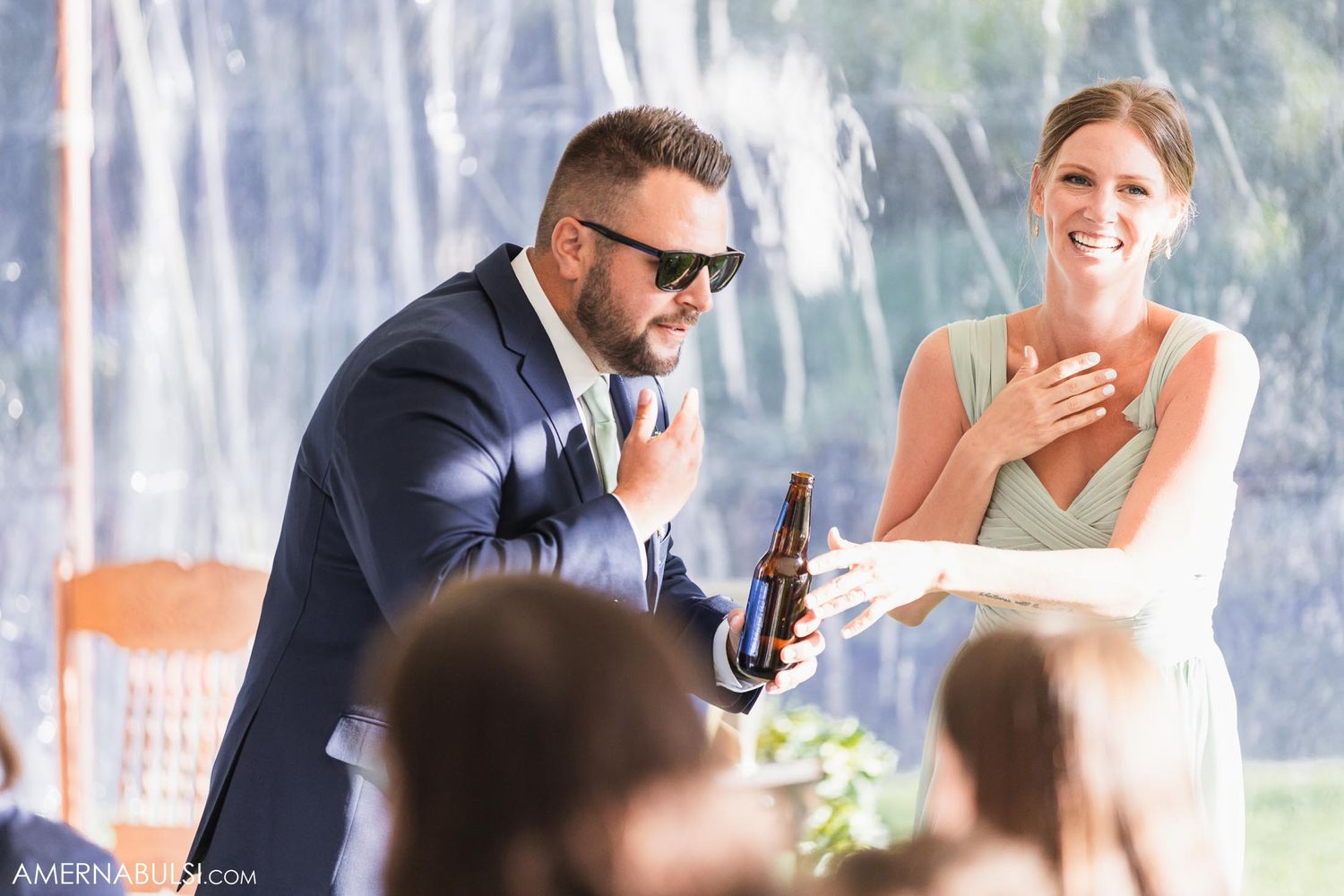 Wedding reception guests enjoying drinks and laughter under white tent.