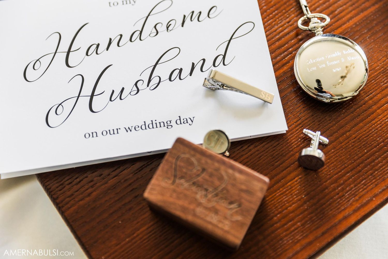 Wedding details flat lay with handwritten card, pocket watch, and accessories on wooden surface.