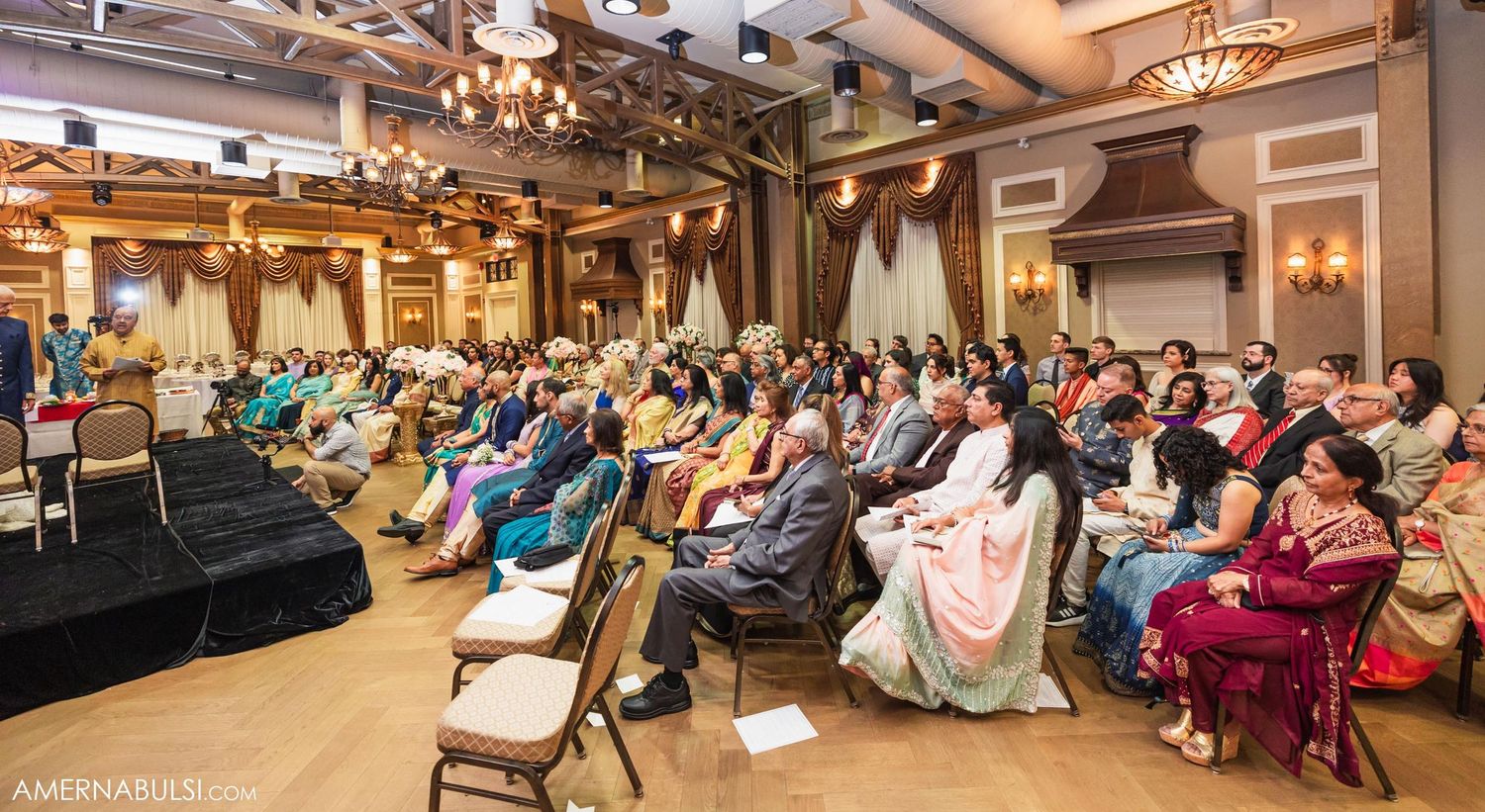 Large crowd gathered in an elegant ballroom for a formal event with ornate lighting and decorative details.