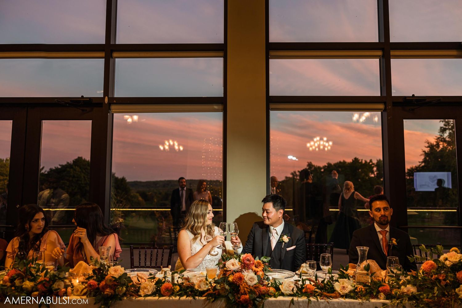 Wedding reception dinner table decorated with floral arrangements and glowing chandeliers at sunset.