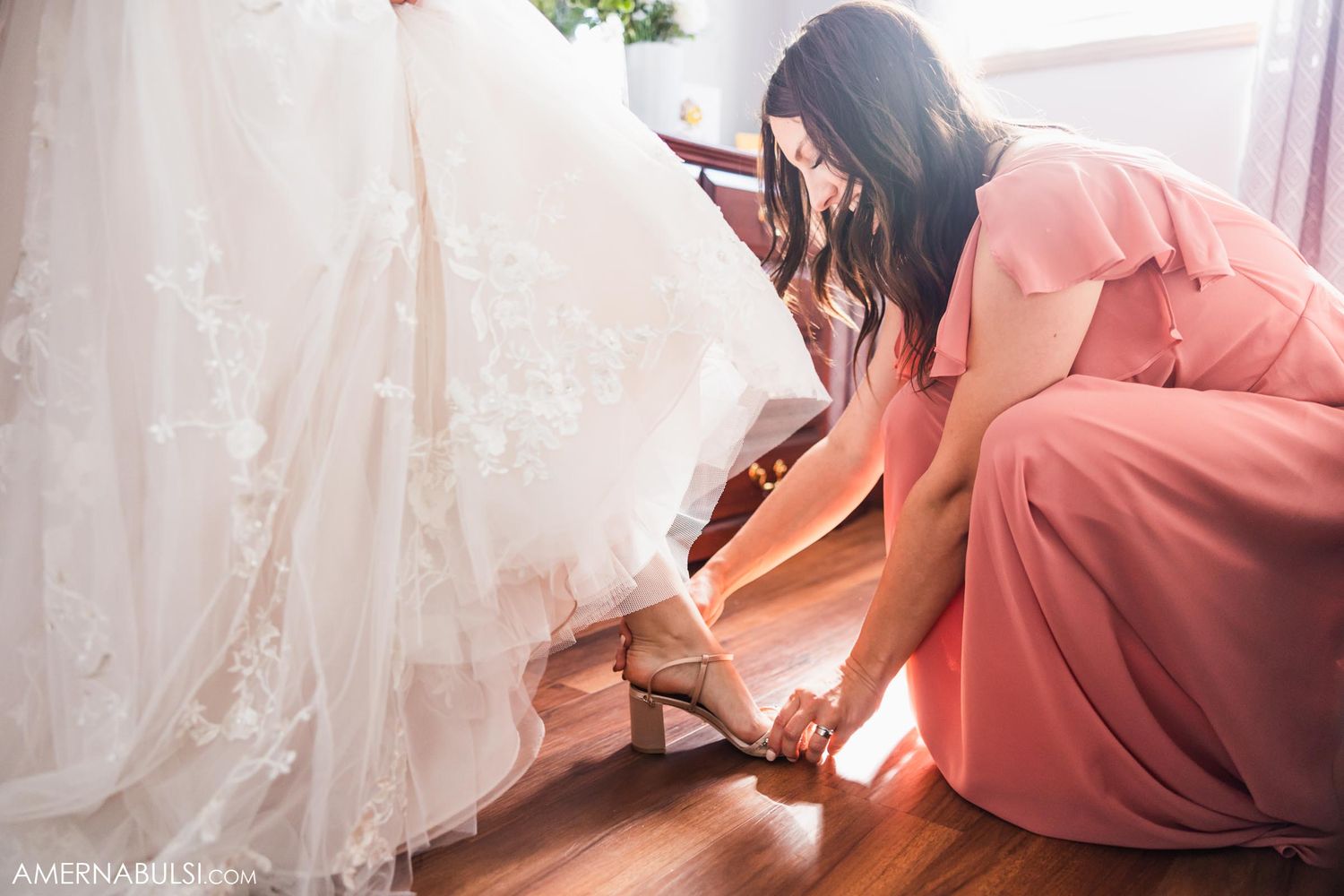 Woman in coral dress helps adjust the hem of a white wedding gown on wooden floor.