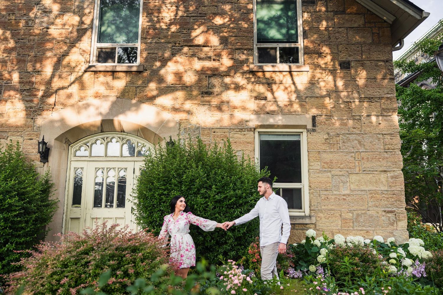 A couple holds hands in front of a stone house with beautiful arched windows and blooming gardens.
