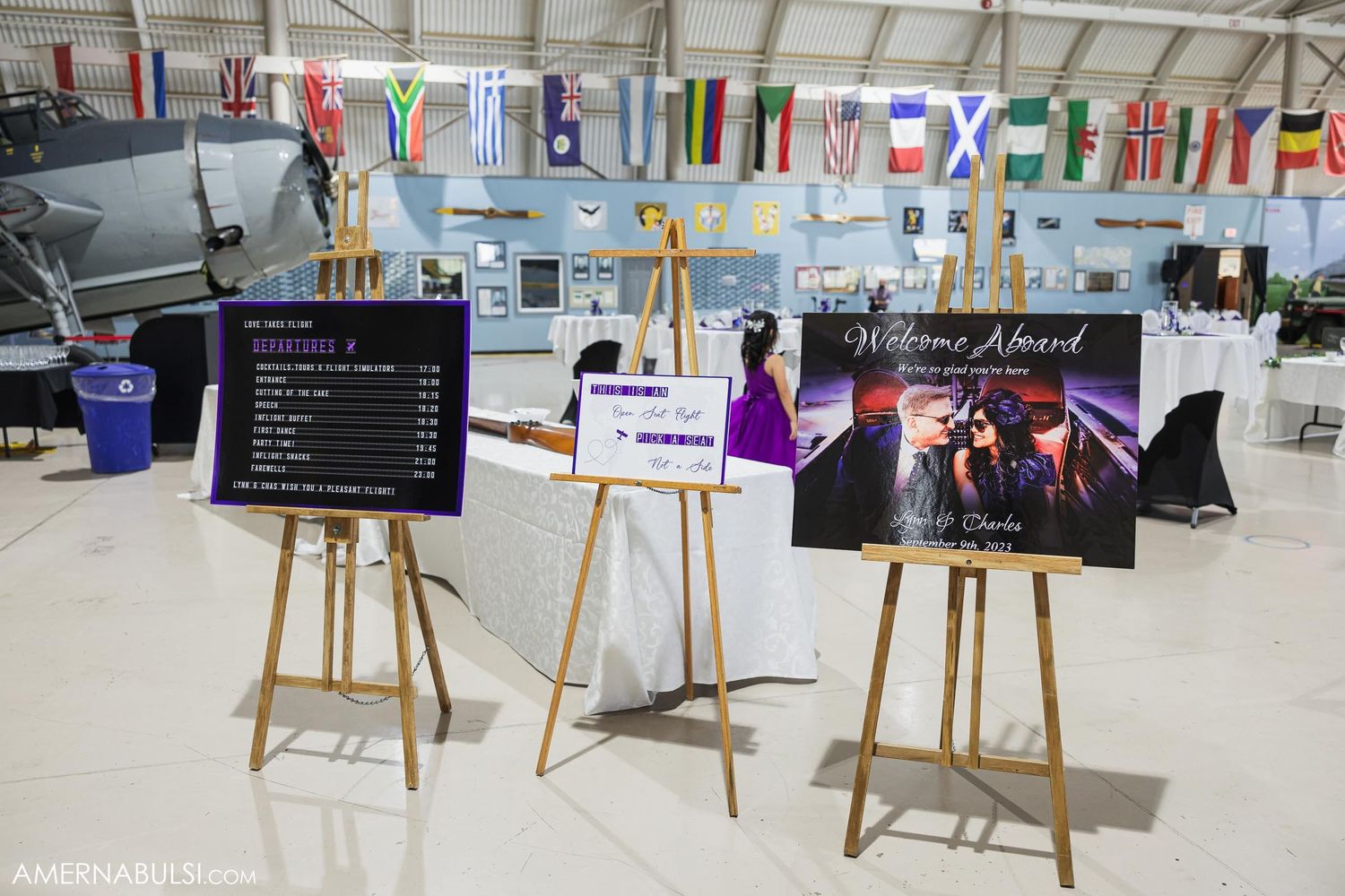 Three wooden easels displaying wedding event signage and photos in front of international flags.