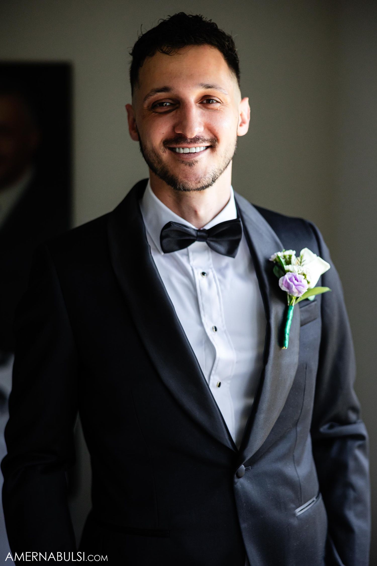 Groom in classic black tuxedo with bowtie and floral boutonniere smiling at camera.