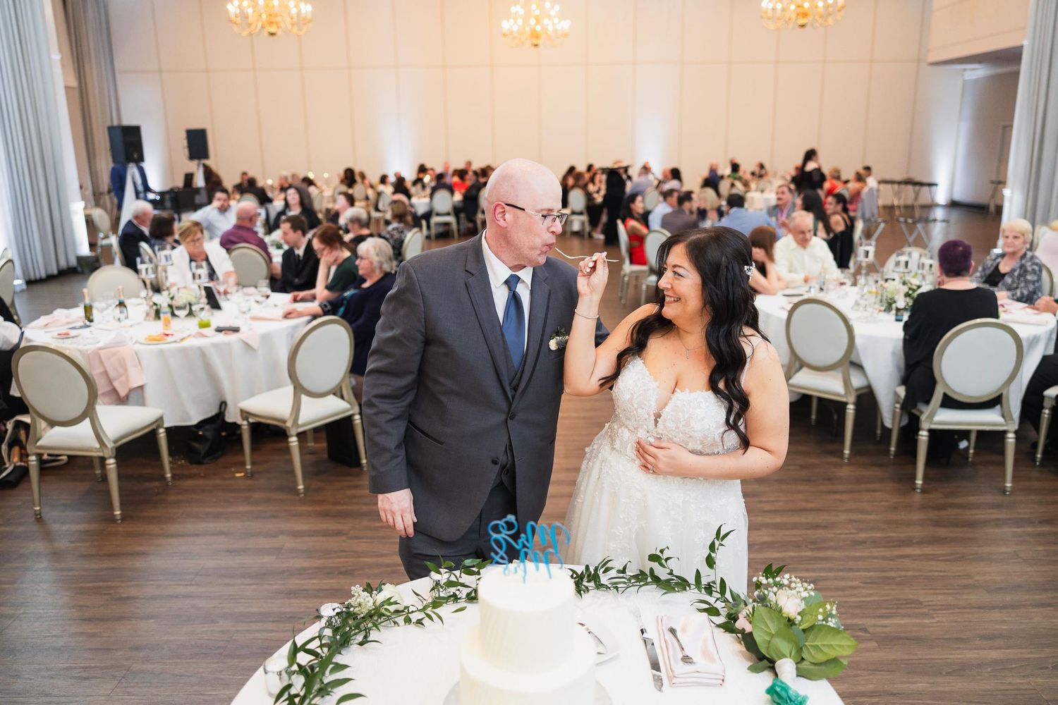 A couple stands behind their wedding cake at an elegant reception with white decor.