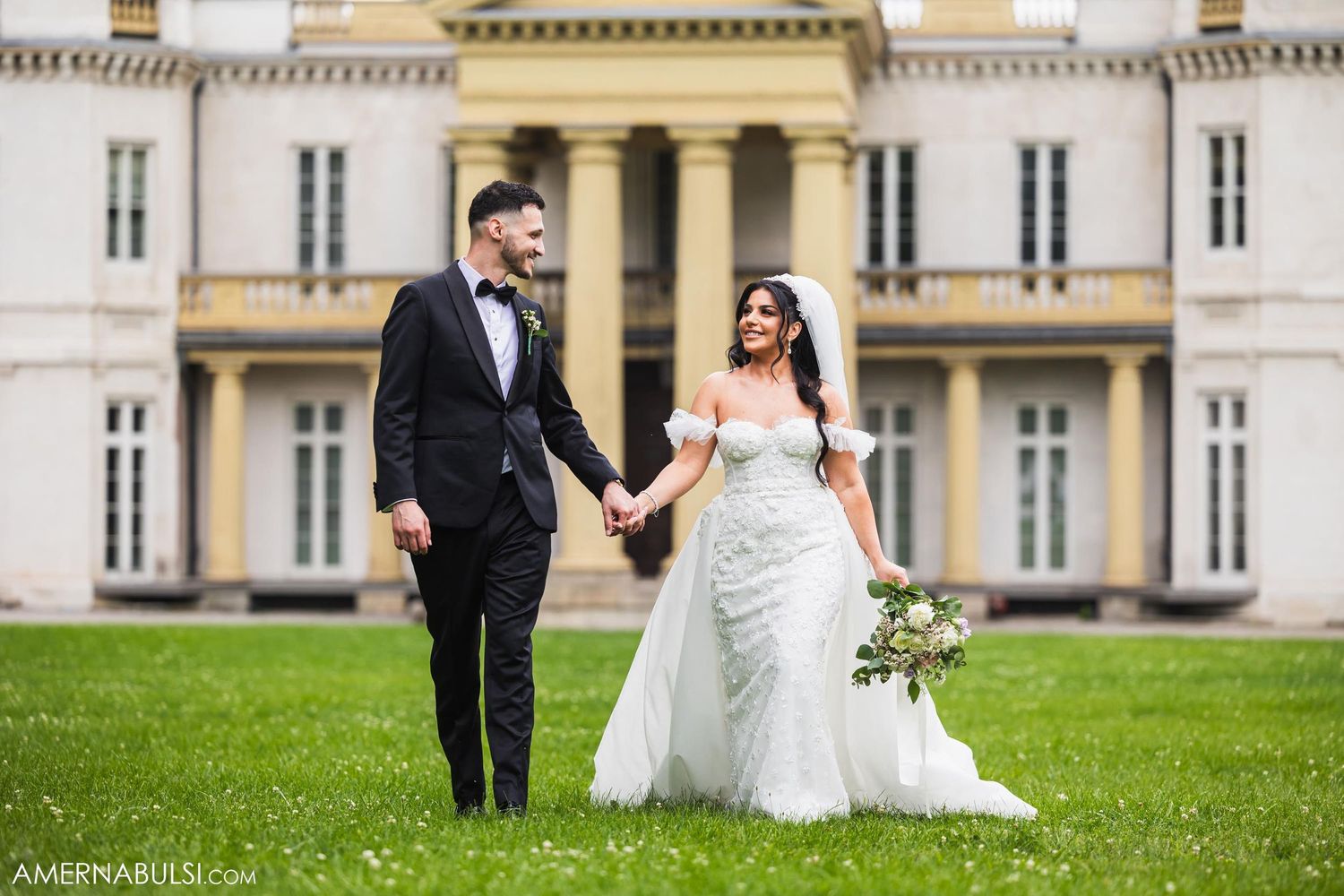 Wedding couple holding hands on the lawn of an elegant mansion with classical architecture.