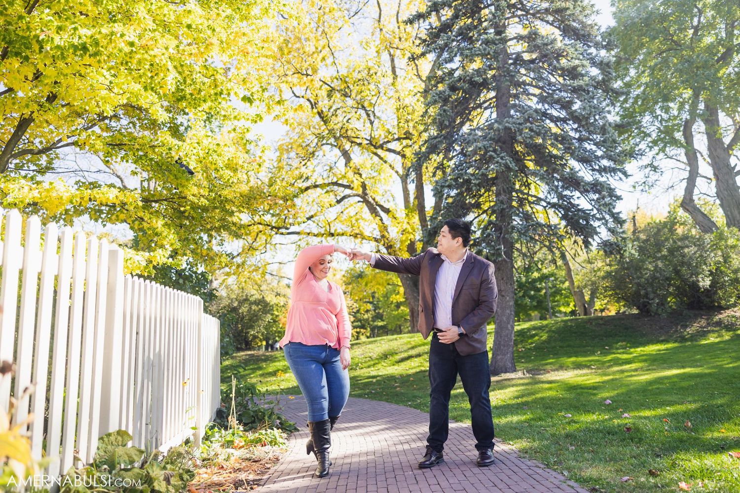 Couple walking along garden path with autumn trees and white picket fence.