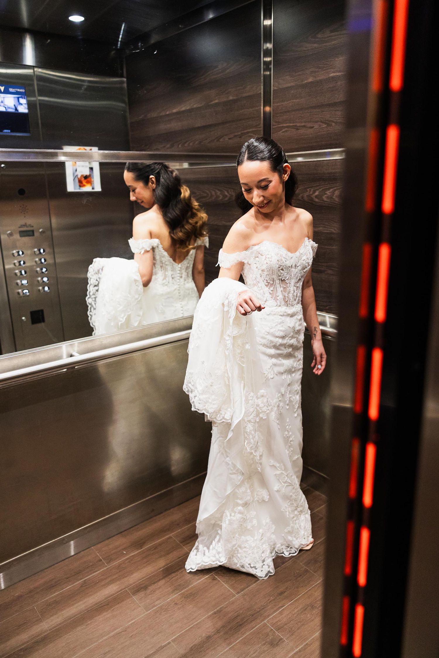 A bride in a lace wedding gown checking her dress in an elevator mirror with wood paneled walls.