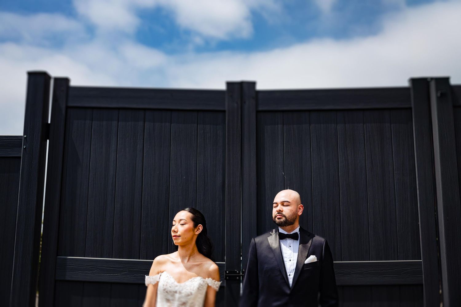 A wedding couple poses dramatically against a black fence under blue skies.