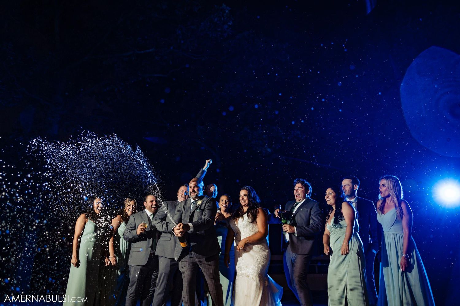 Wedding group cheers under dramatic blue lighting as confetti showers down.