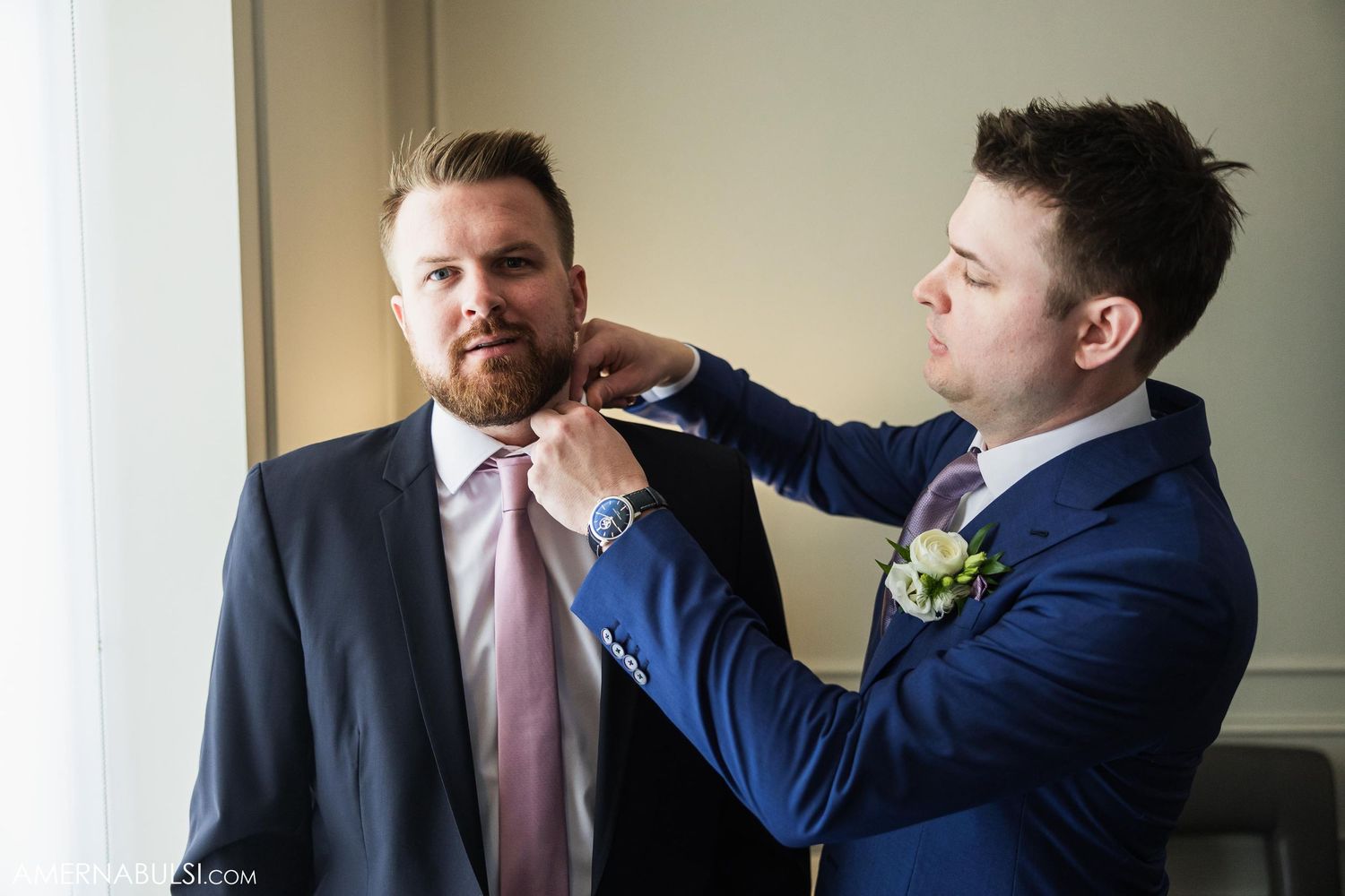 Two people in formal wear helping each other prepare with final tie adjustments.