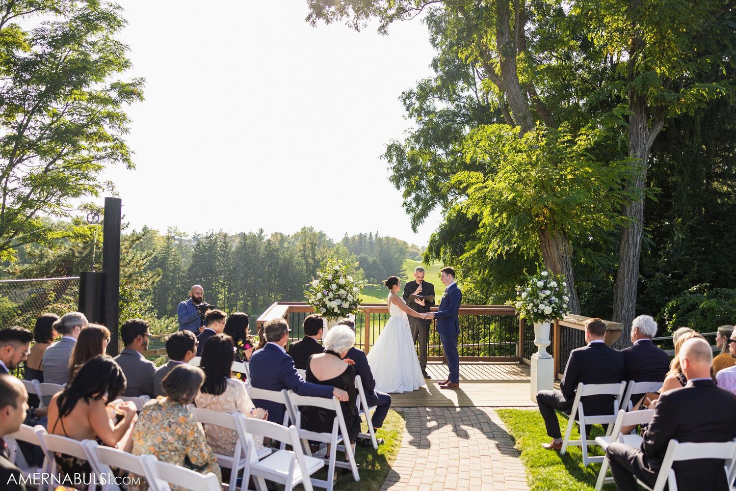 Outdoor wedding ceremony with guests seated on white chairs amid sunlit trees.