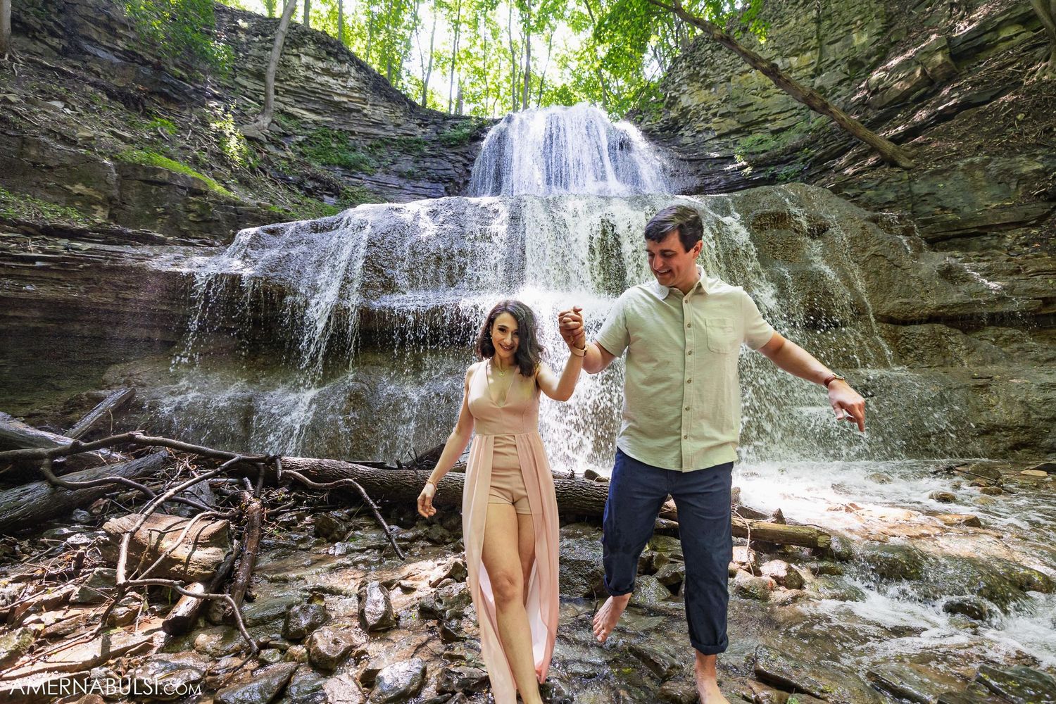 Couple enjoying a playful moment together on rocky terrain in front of a cascading waterfall.