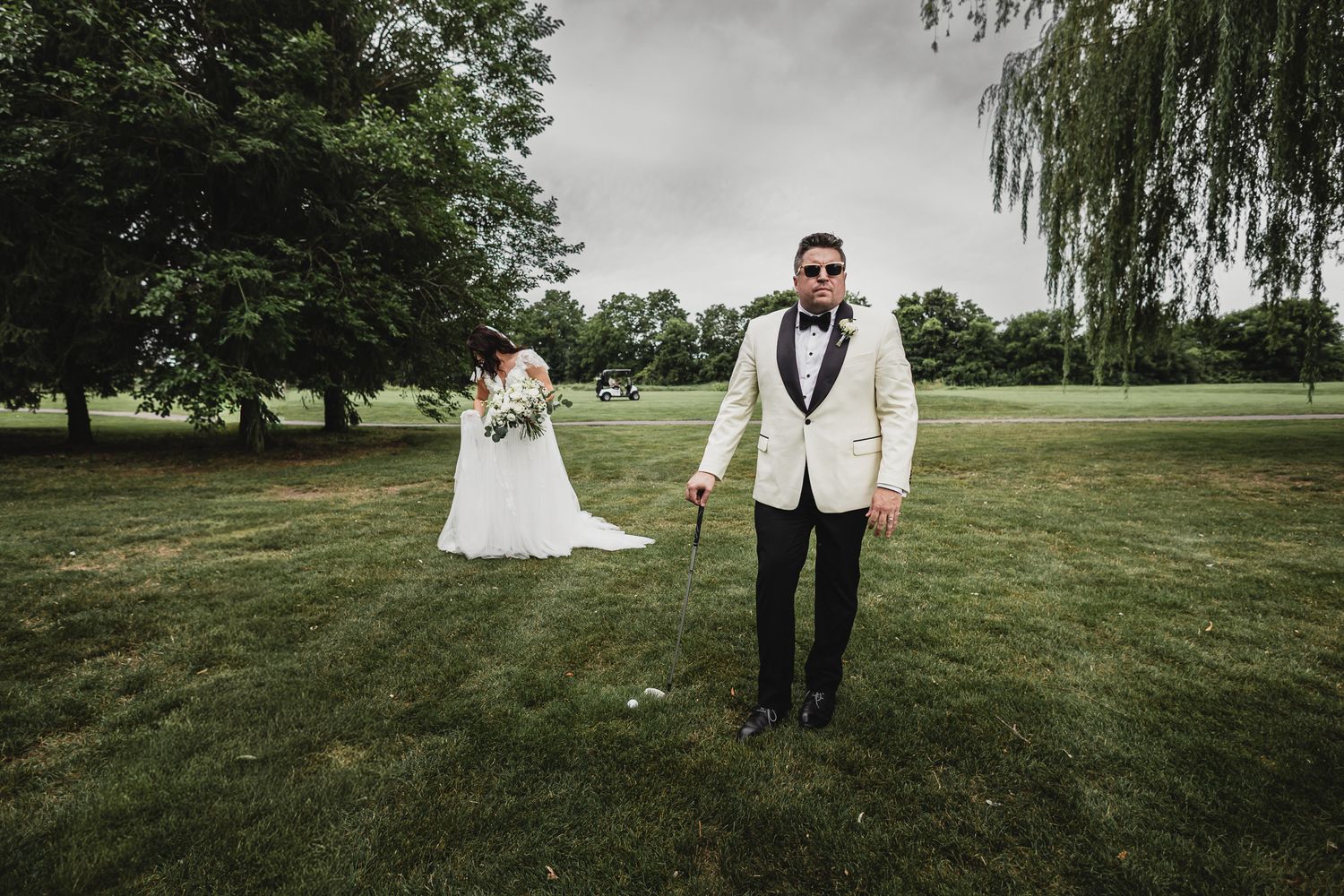 Bride and groom pose on lush green golf course grounds with weeping willow trees in background.