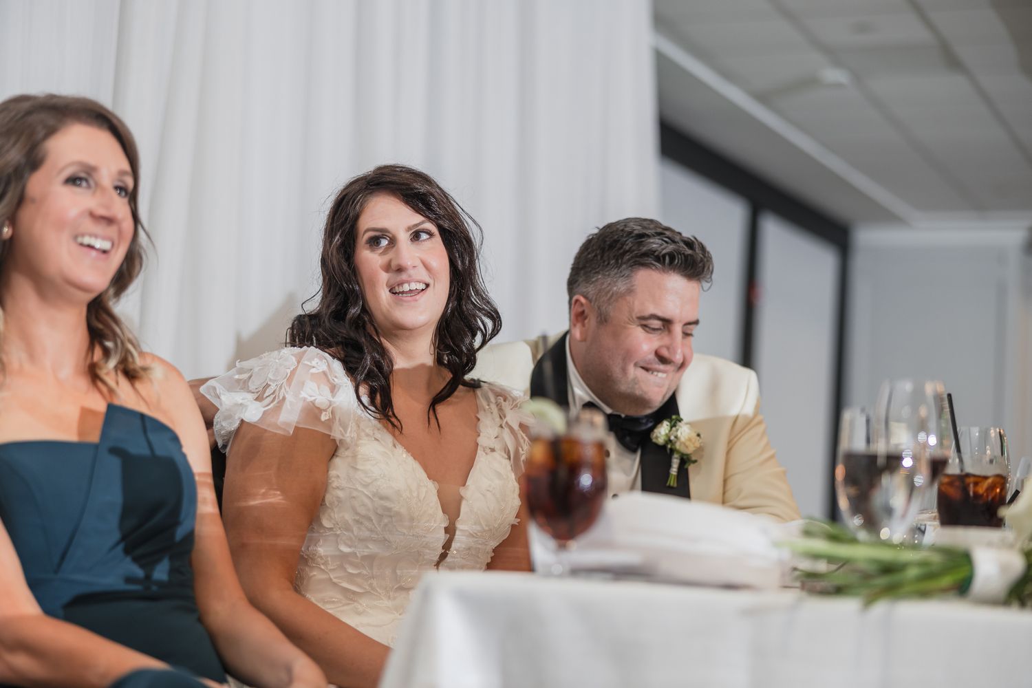 Bride and groom smile at wedding reception head table with bridesmaid in navy blue dress beside them.