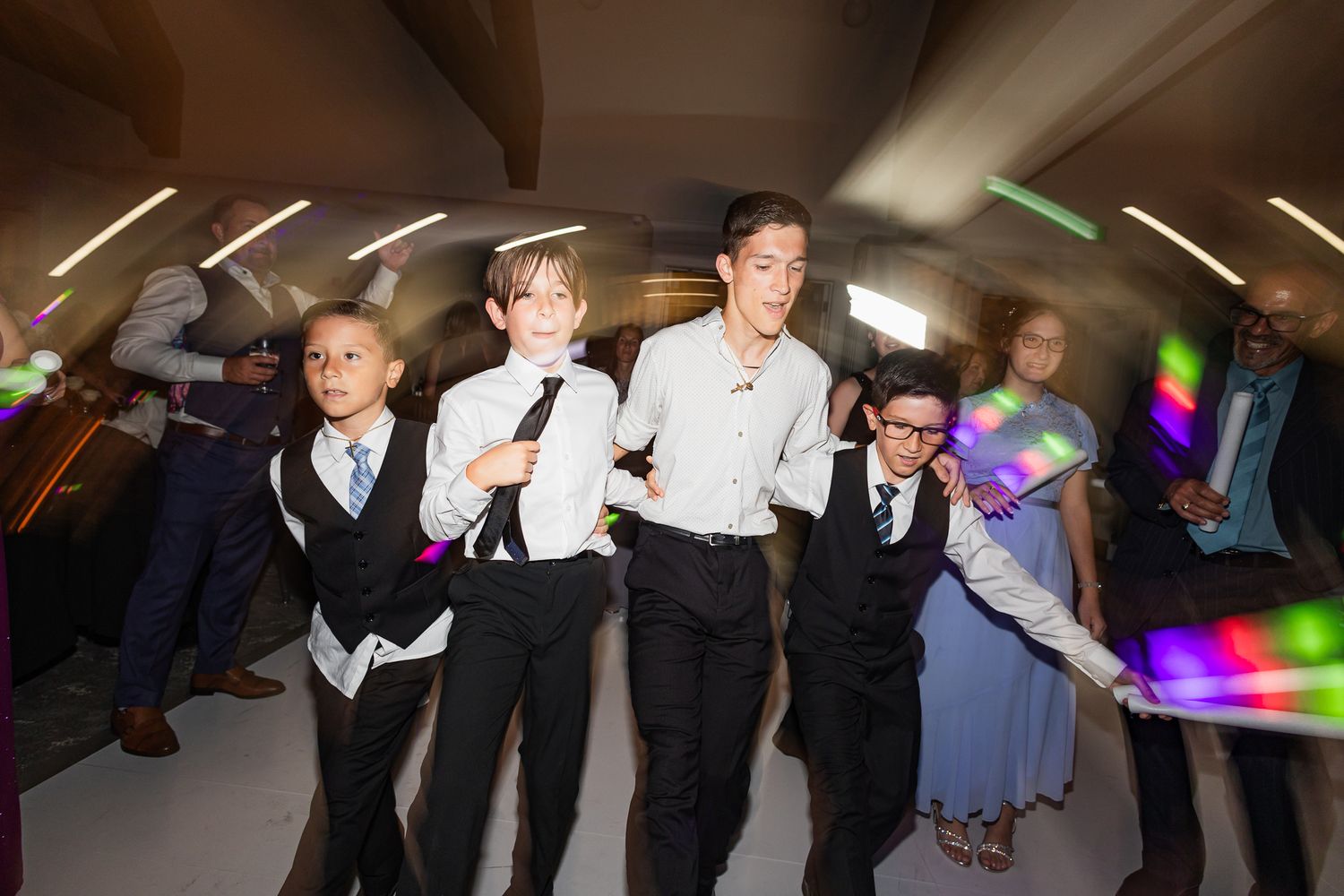 Children and adults in formal attire dance together at a wedding reception on a bright dance floor.