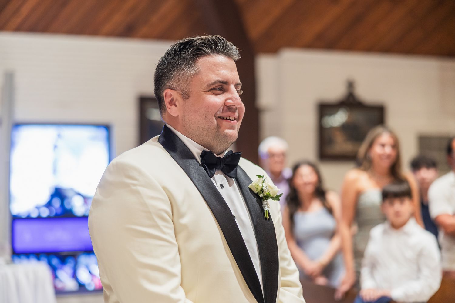 Groom in ivory tuxedo smiles warmly as he watches his bride walk down the aisle.